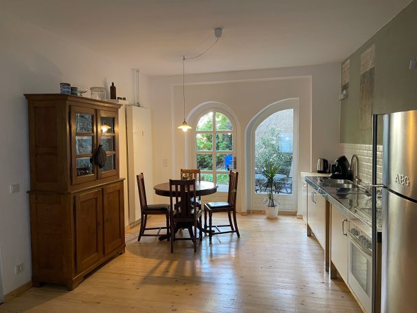 Blick vom Wohnzimmer zur Küche mit Essbereich Helle Küche mit Holzschrank, Tisch, Stühlen und Zugang zu einem Garten.