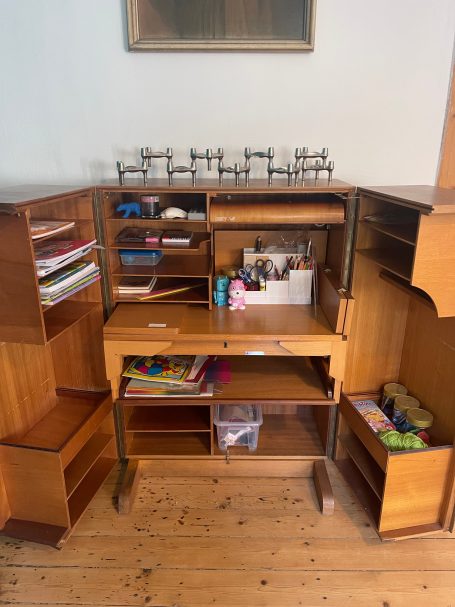 Schreibtisch-Schrank mit Mal- und Bastel-Utensilien Holzkommode mit Schubladen und Regalfächern, gefüllt mit Büchern und bunten Materialien.