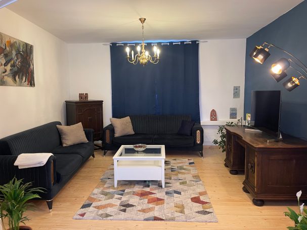 Blick von der Wohnküche ins Wohnzimmer Wohnzimmer mit zwei Sofas, Tisch, Teppich und Fernseher vor blauer Wand. Pflanzen dekorativ platziert.