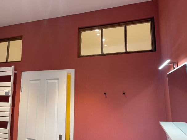 Badezimmer mit Oberlichtern zum Wohnungsflur Wand in rotem Farbton mit einem Fenster und einem weißen Türrahmen.
