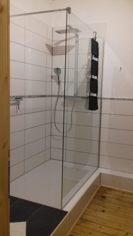 Badezimmer mit großzügiger Regendusche Moderne Dusche mit Glaswand, grauen Fliesen und Holzfußboden.