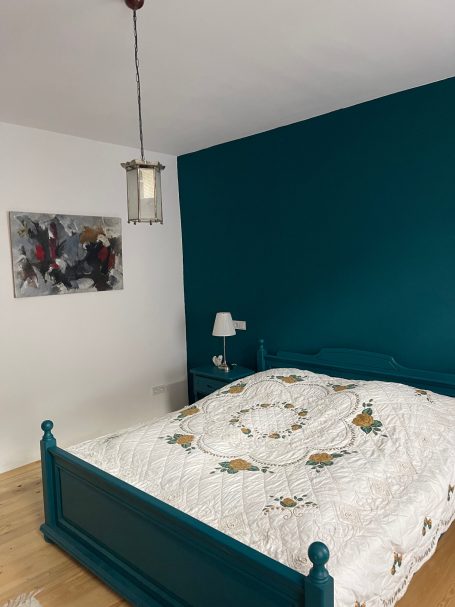 Schlafzimmer mit 180er Vollholz-Bett Schlafzimmer mit grünem Wandakzent, Bett mit floralem Bettzeug und moderner Wandkunst.