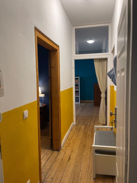 Wohnungseingang Ein schmaler Flur mit gelben Wänden, Holzboden und Zugang zu einem Raum mit Vorhang.