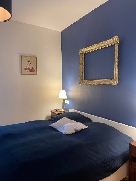 Schlafzimmer mit 180er Bett Dunkelblau gestrichene Wand hinter einem Bett mit einer weißen Bettdecke und einem Nachttisch.