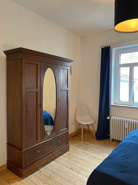 Schlafzimmer mit Kleiderschrank und Spiegel Holzschrank mit Spiegel, blauer Bettwäsche und einem weißen Stuhl in einem gemütlichen Raum.